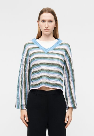Femme portant un pull en maille ample rayé aux couleurs pastel et col en V sur une chemise blanche, associé à un pantalon noir taille haute.