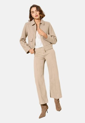 Mujer con chaqueta beige, pantalones de pierna ancha de talle alto a juego, camiseta blanca y botines de ante marrón con tacón alto, de pie contra un fondo liso.