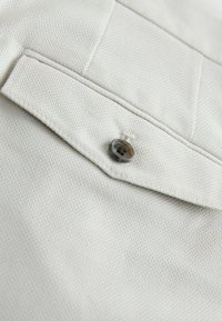 Gros plan sur un tissu beige clair texturé avec une poche à rabat cousue, sécurisée par un seul bouton foncé.