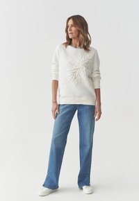 Felpa bianca con design a forma di foglia tridimensionale testurizzato sul davanti. Abbinata a jeans azzurri e sneakers bianche. Tessuto morbido, vestibilità comoda.