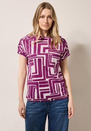 T-shirt court violet et blanc à manches courtes avec des motifs géométriques. Tissu doux avec une coupe ample et un col rond, associé à un jean bleu.