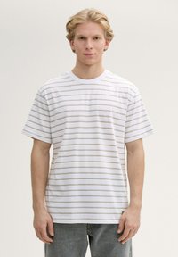 Camiseta de algodón blanca con delgadas rayas horizontales verdes, cuello redondo, mangas cortas y un corte relajado. El modelo lleva unos jeans azul claro.