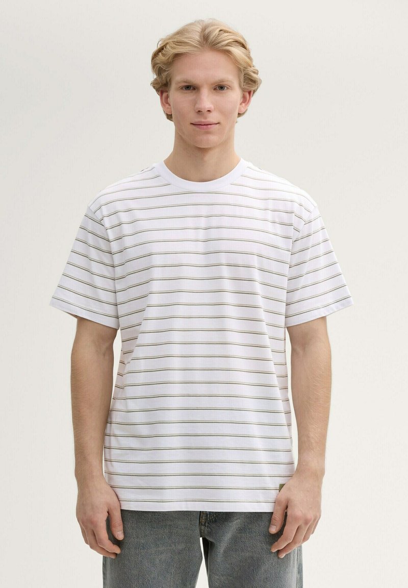 Camiseta de algodón blanca con delgadas rayas horizontales verdes, cuello redondo, mangas cortas y un corte relajado. El modelo lleva unos jeans azul claro.