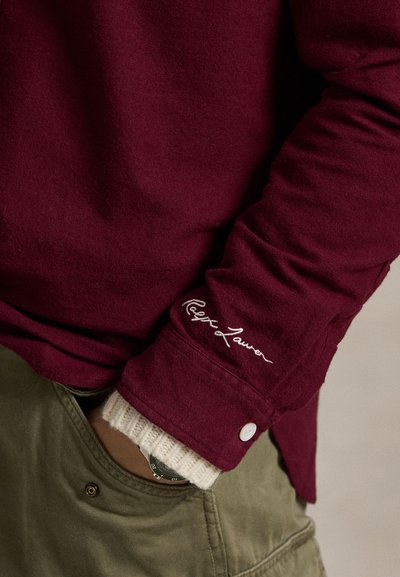 Μακρύ μανίκι μπορντώ πουκάμισο με ριγέ υφή, διαθέτοντας λευκό κεντητό λογότυπο στην μανσέτα και αντίθετη μπεζ μανσέτα τύπου βαρέλι.