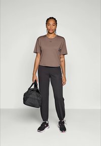 Brun cropped t-skjorte med korte ermer og en logo, kombinert med svarte joggebukser. Svart treningsbag og sportssko med rosa detaljer.