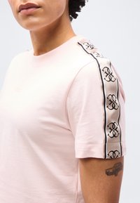 Άτομο που φοράει ροζ ανοιχτού χρώματος κοντομάνικο πουκάμισο με φαρδιά ρίγα σε μπεζ και μαύρο στο ύψος του ώμου και μικρό τατουάζ αυτοκινήτου στον πάνω βραχίονα.