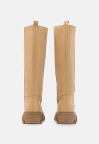 Vysoké béžové kožené čižmy s hrubými, robustnými gumovými podrážkami, zobrazené zozadu na jednoduchom bielom pozadí.