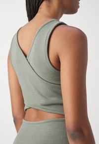 Olivgrön ärmlös topp i texturerat tyg. Har en korsad design på ryggen och en kort skärning som framhäver en åtsittande siluett.