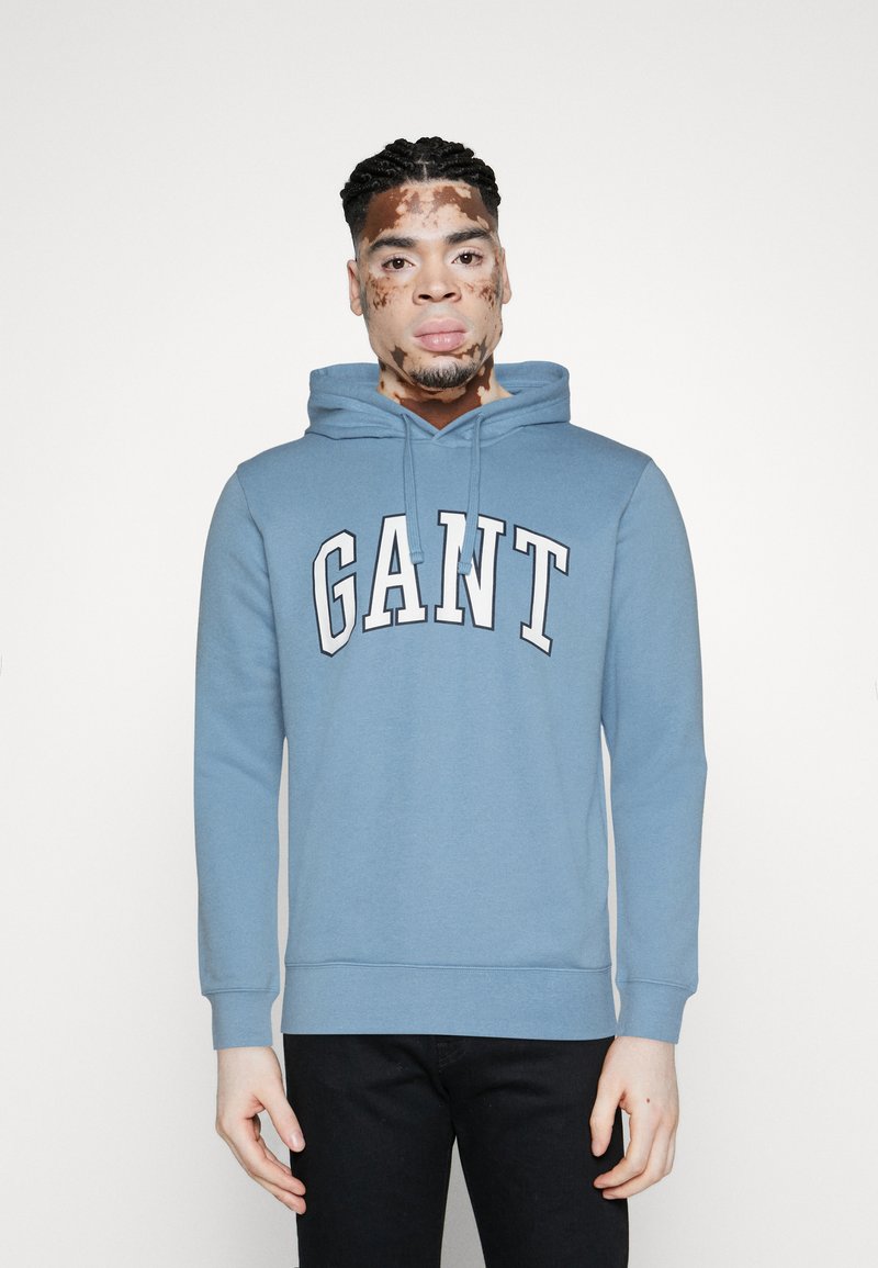 Felpa GANT azzurro chiaro con tasca a marsupio frontale, cappuccio con cordino e grande lettering bianco. Tessuto morbido con polsini a costine.
