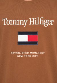 Rjava bombažna T-majica z belim vezenim logotipom "Tommy Hilfiger", barvnim zastavnatim dizajnom v mornarsko modri, rdeči in beli barvi, z napisom "USTANOVLJENO MCMLXXXV NEW YORK CITY" spodaj.