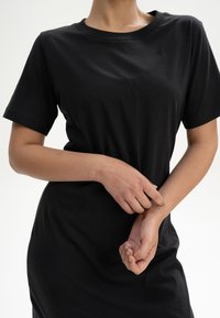 Schwarzes Baumwoll-T-Shirt mit kurzen Ärmeln, rundem Ausschnitt und lässiger Passform. Der Stoff wirkt glatt mit subtiler Textur und wird lässig getragen.