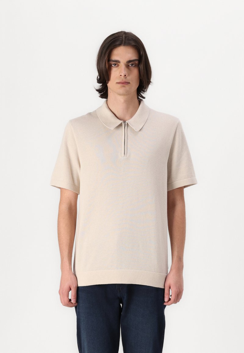 Beige strikket polo med glidelås i halsen, korte ermer og løs passform. Myk tekstur og minimalistisk design. Kombinert med mørke denimjeans.