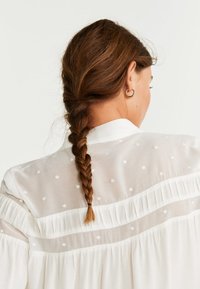 Weiße Bluse mit einem strukturierten Muster, ausgestattet mit einem geflochtenen Rückendesign und transparenter Stoff mit bestickten Punkten, sowie einem gesch gathered Rüschendetail.