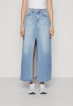 Person iført en lyseblå denim maxi nederdel med en frontslids, hvide sneakers og en hvid ærmeløs top, stående mod en ensfarvet baggrund.