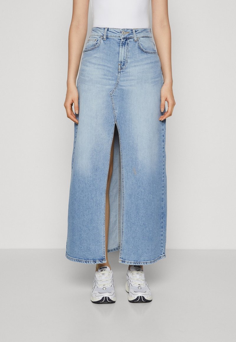 Personne portant une jupe maxi en denim bleu clair avec une fente avant, des baskets blanches et un haut blanc sans manches, debout devant un fond uni.