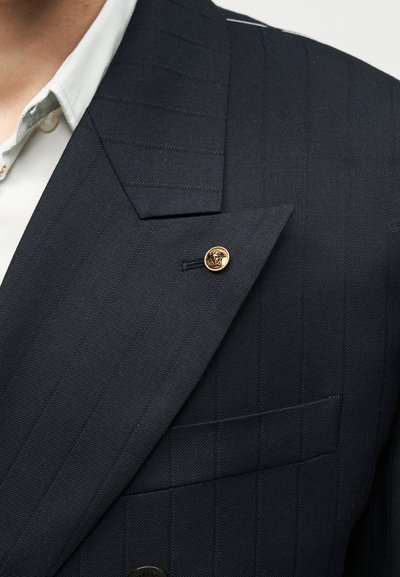 Σακάκι ναυτικού με ρίγες και καρφίτσα με χρυσό κεφάλι Μέδουσας στο πέτο, φορεμένο πάνω από γιακά λευκού πουκαμίσου.