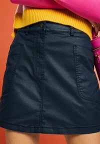 Ciemnoniebieska spódnica z ekoskóry z dwoma przednimi kieszeniami, widocznymi szwami i paskiem zapinanym na guzik, w połączeniu z żółtym swetrem.