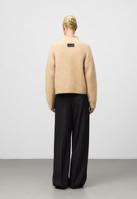 Pull en mélange de laine beige avec un col montant, un ourlet côtelé et une étiquette noire rectangulaire au dos, associé à un pantalon noir ample.
