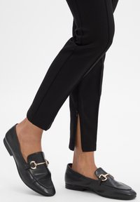 Beine überkreuzt, schmal geschnittene schwarze Hosen mit Reißverschlüssen an den Knöcheln und schwarze Lederloafers mit goldfarbenem Metallzierteil.