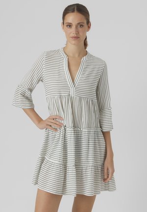 Vestido blanco con rayas verticales negras, con cuello cuadrado, mangas tres cuartos y un dobladillo con volantes en capas. Hecho de una tela ligera.