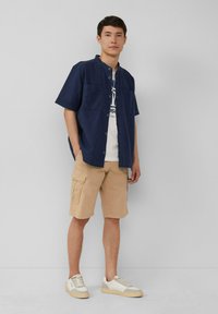 Marineblaues Kurzarmhemd über einem weißen Grafik-T-Shirt, kombiniert mit beigen Cargo-Shorts und hellen Turnschuhen mit cremefarbenen Akzenten.