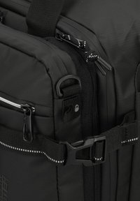 Zaino nero realizzato in materiale resistente, con scomparti con zip, tracolla regolabile e un dettaglio con fibbia piatta impreziosita da cuciture bianche.