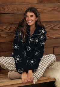 Sort fleece-pullover med hvid blomstret broderi, høj hals og lynlåslukning. Kombineret med ternede pyjamasbukser og beige hjemmesko. Træbaggrund.