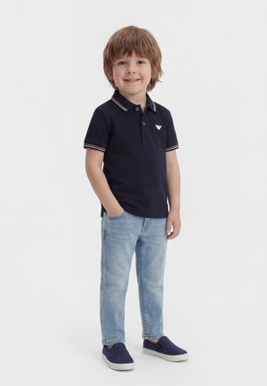 Jeune garçon souriant, debout avec une main dans la poche, portant un polo bleu marine, un jean bleu clair et des chaussures à enfiler bleu marine sur fond blanc.