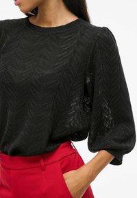 Blouse noire texturée avec un motif en chevrons, col rond et manches légèrement volumineuses, associée à un pantalon rouge taille haute.