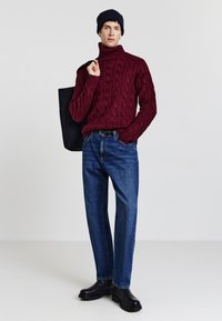 Burgunderfarbener, kabelgestrickter Rollkragenpullover, blaue Jeans, schwarze Stiefel und eine schwarze Tragetasche. Einheitliche Farbe, strukturierte Muster, lockere Passform.