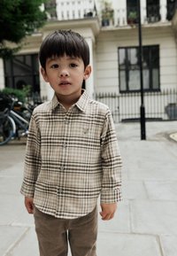 Camicia a quadri beige e marrone con maniche lunghe e bottoni, abbinata a pantaloni di velluto a coste marroni. Ambientazione urbana all'aperto con biciclette sullo sfondo.