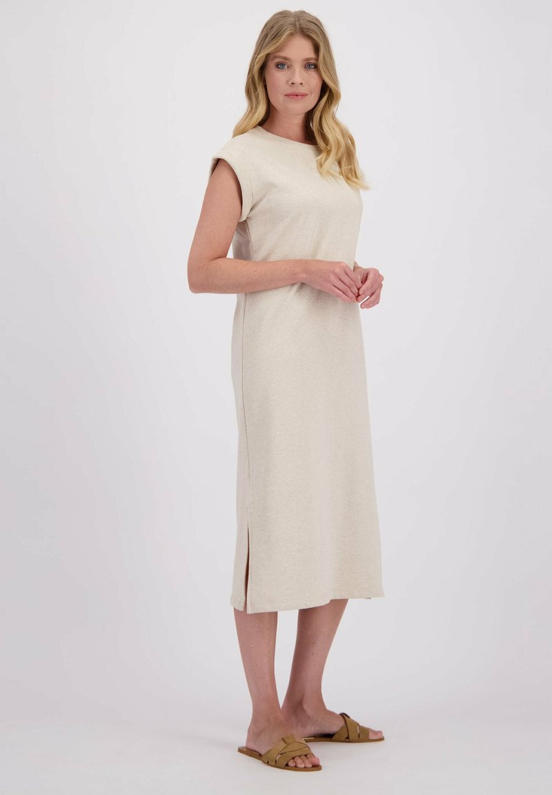 Femme aux longs cheveux blonds portant une robe midi sans manches beige et des sandales à semelle plate tan, se tenant devant un fond clair et uni.