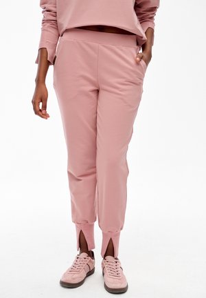 Kobieta w jasnoróżowych spodniach dresowych z rozcięciami przy kostkach i pasujących sneakersach, stojąca z jedną ręką w kieszeni.