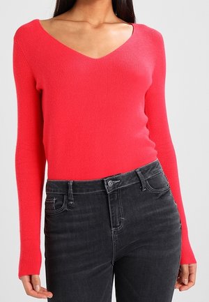 Svetr s výstřihem do V, pletený v jemných letech, v jasně korálové barvě, s dlouhými rukávy. Nosený s tmavě šedými džíny, které zvýrazňují ladnou siluetu.