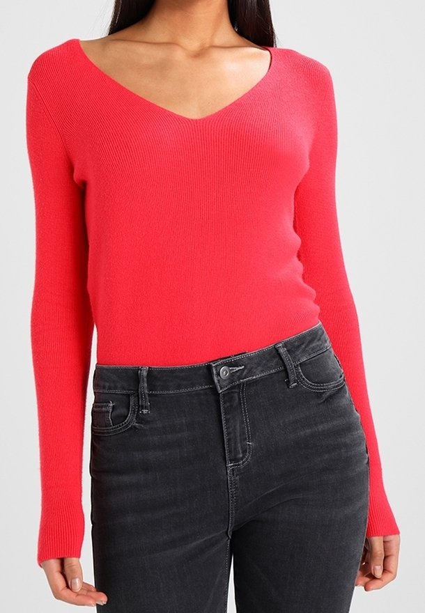Sweter z dekoltem w serek, dzianinowy, w jasnym koralowym kolorze, z długimi rękawami. Noszony z ciemnoszarymi dżinsami, podkreślający dopasowaną sylwetkę.