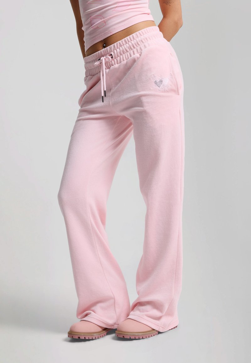 Kvinde iført lyserøde bukser med vide ben og hjertebroderi på lommen, matchende lyserøde sko og en cropped top mod en ensfarvet baggrund.