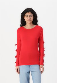 Rød langermet skjorte med teksturerte sløyfer ved mansjettene, rund halsutskjæring og en tettsittende design, kombinert med lyseblå jeans.