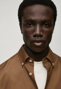 Brown button-up shirt with a smooth texture, featuring white buttons and a collar, worn over a white T-shirt. Close-up portrait.