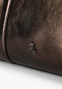 Bolso de cuero marrón texturizado con un pequeño emblema de calavera y huesos cruzados en negro. Acabado suave con un brillo metálico. Diseño compacto.