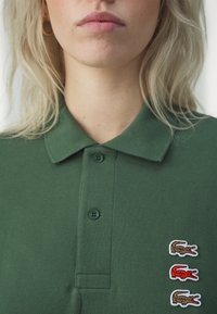 Zelený polo tričko s texturovanou tkaninou, tři vyšívané loga krokodýla v hnědé a červené barvě na levé hrudi a klasický design límečku.