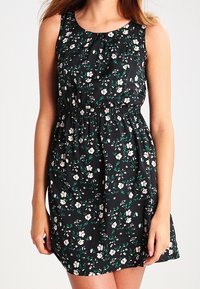 Vestido floral con fondo negro, que presenta flores blancas y verdes, diseño sin mangas, cintura elástica y textura de tela suave.