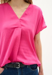 Blusa rosa a maniche corte con scollo a V e vestibilità rilassata. Indossata con una cintura nera e jeans in denim blu. Tessuto dalla consistenza liscia.