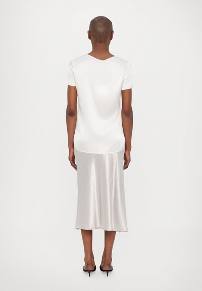 Pessoa com cabelo curto, vestindo uma blusa branca de manga curta, uma saia midi de cetim prateada e mules pretas de salto, em pé contra um fundo branco.