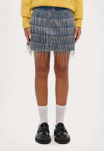 Denimrock mit Fransenverzierungen aus silberartigen Perlen, mit einem Taillengummi und einer Passform, die über die Knie reicht. Schwarze Slipper und weiße Socken.