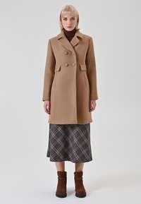 Brauner Wollmantel mit doppelreihiger Knopfleiste, großen Knöpfen und einem Reverskragen, getragen über einem karierten Rock und braunen Stiefeletten.