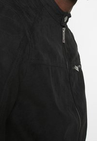 Chaqueta de cuero negra con una textura suave, que presenta un cuello alto, cremalleras plateadas y costuras minimalistas a lo largo de las uniones.