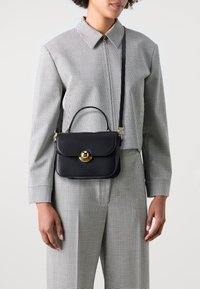 Čierna kožená kabelka so zlatým kruhovým uzáverom, obdĺžnikový tvar, tenký ramenný popruh a horné madlo. Modelka má na sebe kockovaný outfit.