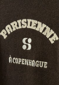 Hnedý pletený textil s vyšívaným bielym textom: "PARISIENNE" oblúkom nad "à COPENHAGUE" a s kruhovým logom s dvoma bodkami.
