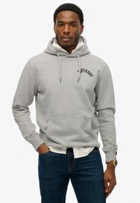 Grijze hoodie met trekkoorden en een voorkeurszak, met de tekst "Superdry". Draag het over een witte shirt, gecombineerd met donkerblauwe jeans.