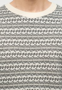 Κοντινό πλάνο ενός ατόμου που φοράει πλεκτό πουλόβερ με ασπρόμαυρο μοτίβο και λαιμόκοψη σε κρεμ απόχρωση.